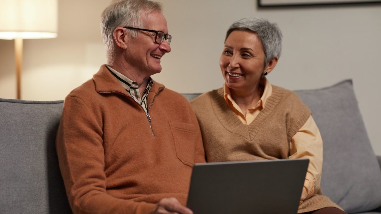 older couple smiling at each other and sitting on couch with laptop
