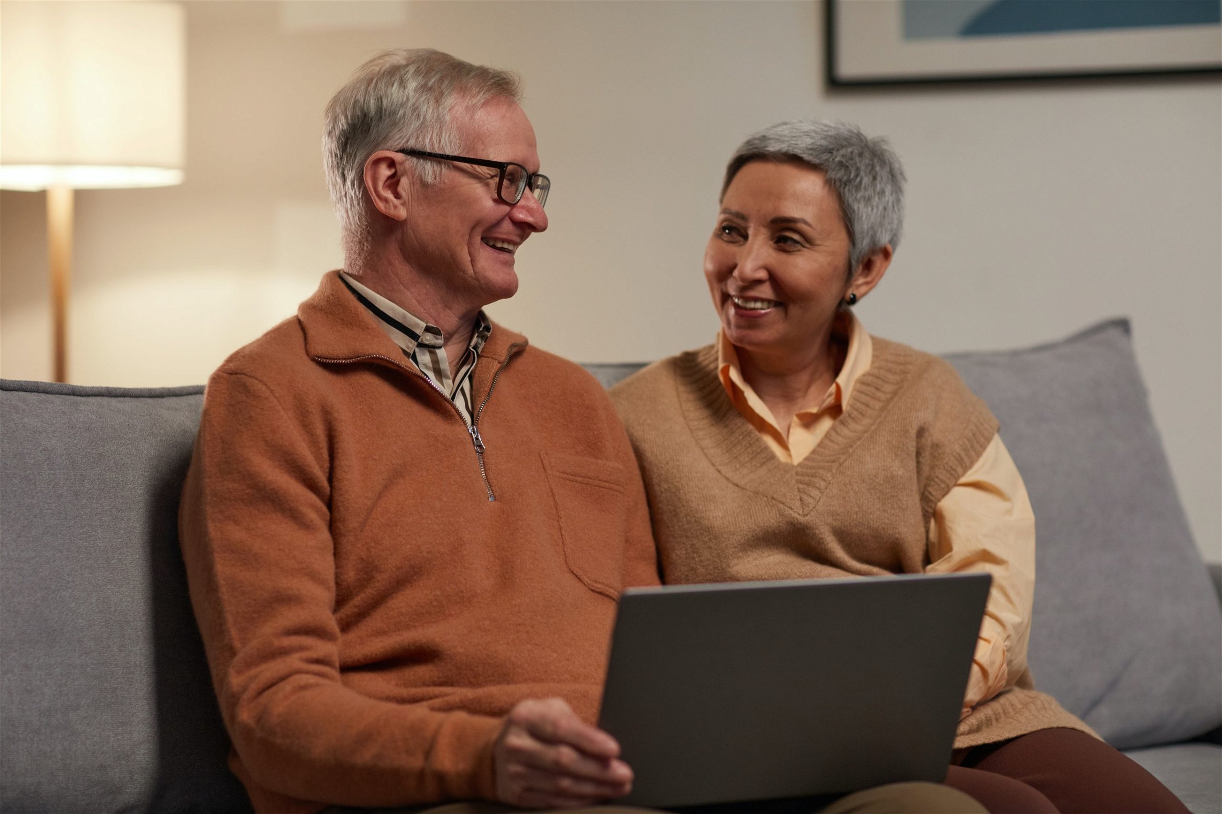 older couple smiling at each other and sitting on couch with laptop