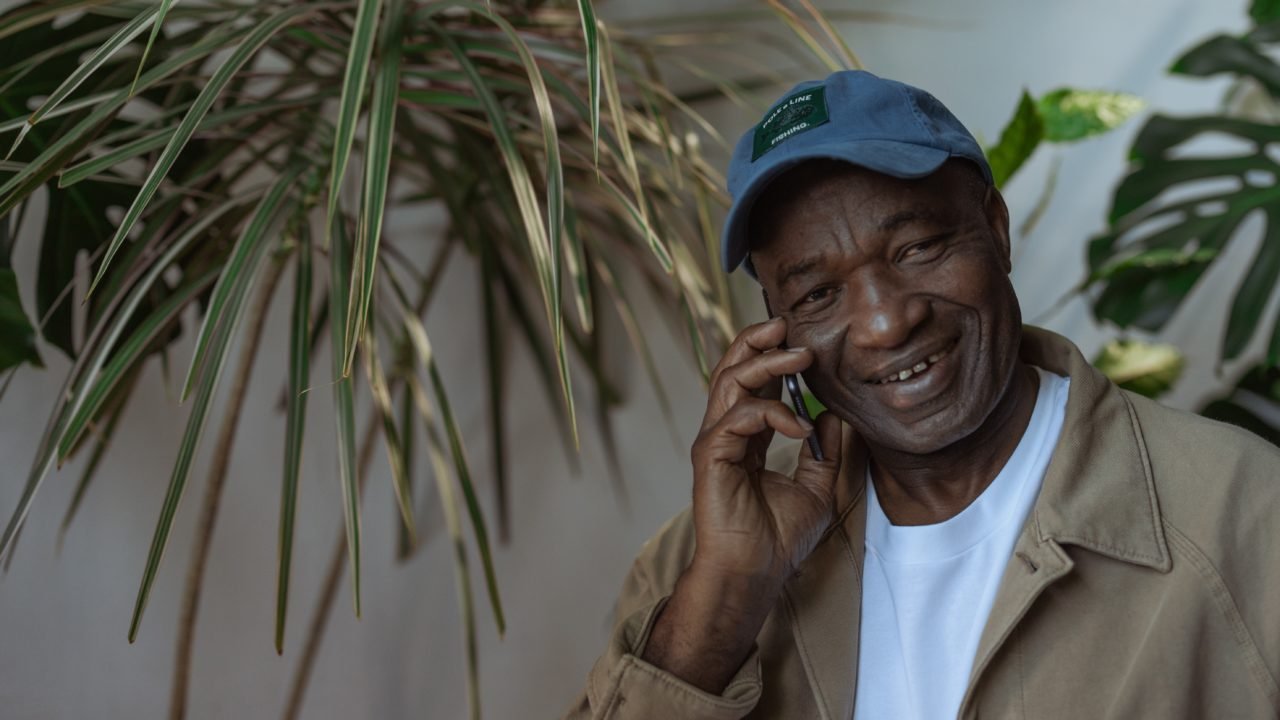 A man wearing a baseball cap smiles while on a phone call.