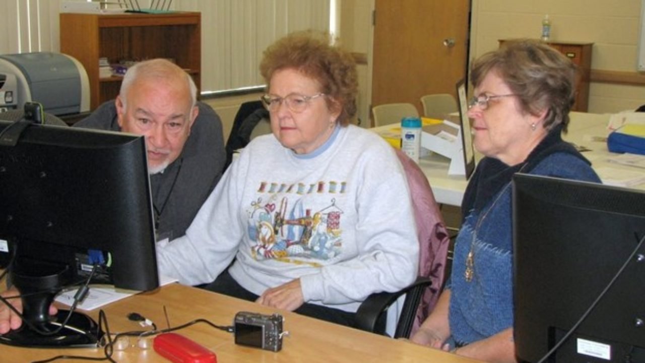 A group of three older adults gather around a desktop computer. They look intently at the screen.