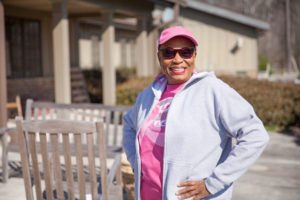 A woman in a pink shirt and a hoodie stands facing the camera with her hands on her hips. She is smiling and the day is sunny. 