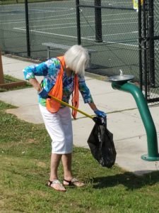 A woman wearing an orange vest uses a grabber to pick up trash in a park and put it in a trashbag. 